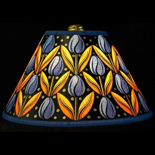 Hand painted small 10” paper empire lampshade covered in playful Rows of blue tulips contrasted by yellow shoots on a black background with gold dots on top and a bold blue trim  lit up, bringing warmth to any room